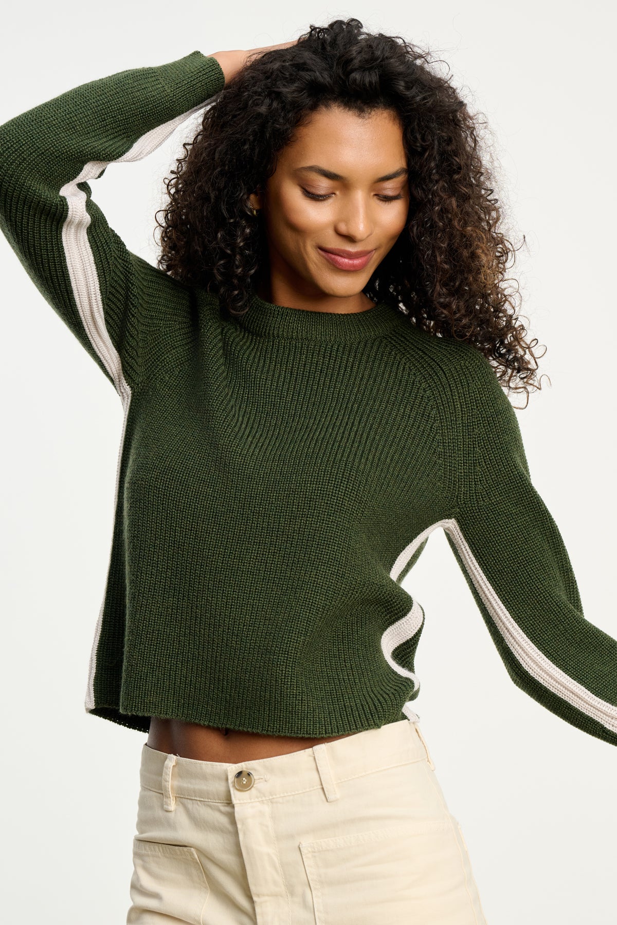 A woman with curly hair wears the SAVANNAH SWEATER by Velvet by Graham & Spencer—a green wool blend crewneck with white striped sleeves—and beige pants, standing with one arm raised.-39800362467521