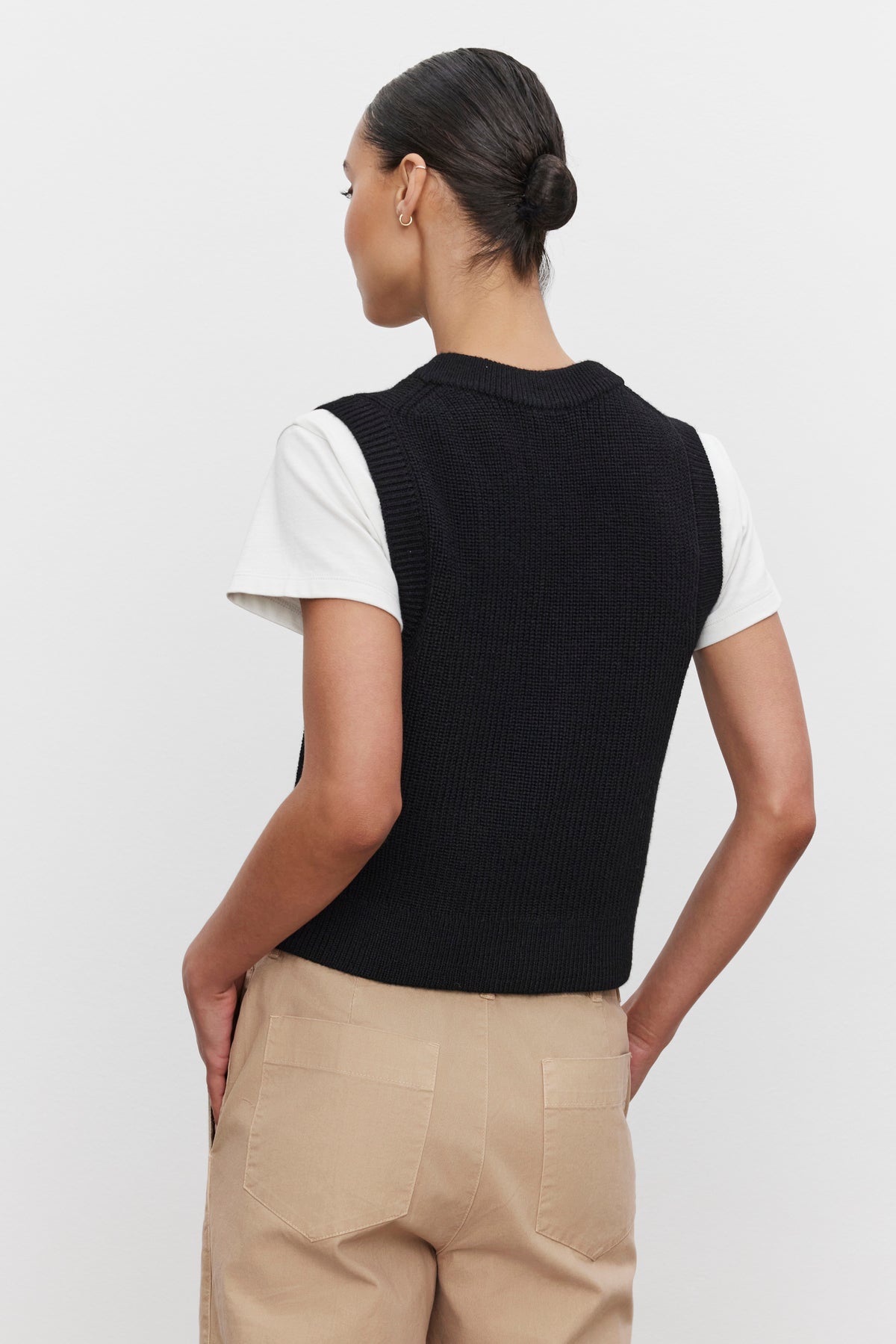   Rear view of a person with hair in a bun, wearing the LU SWEATER VEST by Velvet by Graham & Spencer. The vest is black, sleeveless, and features a v-neckline. It is layered over a white T-shirt and paired with beige pants. 