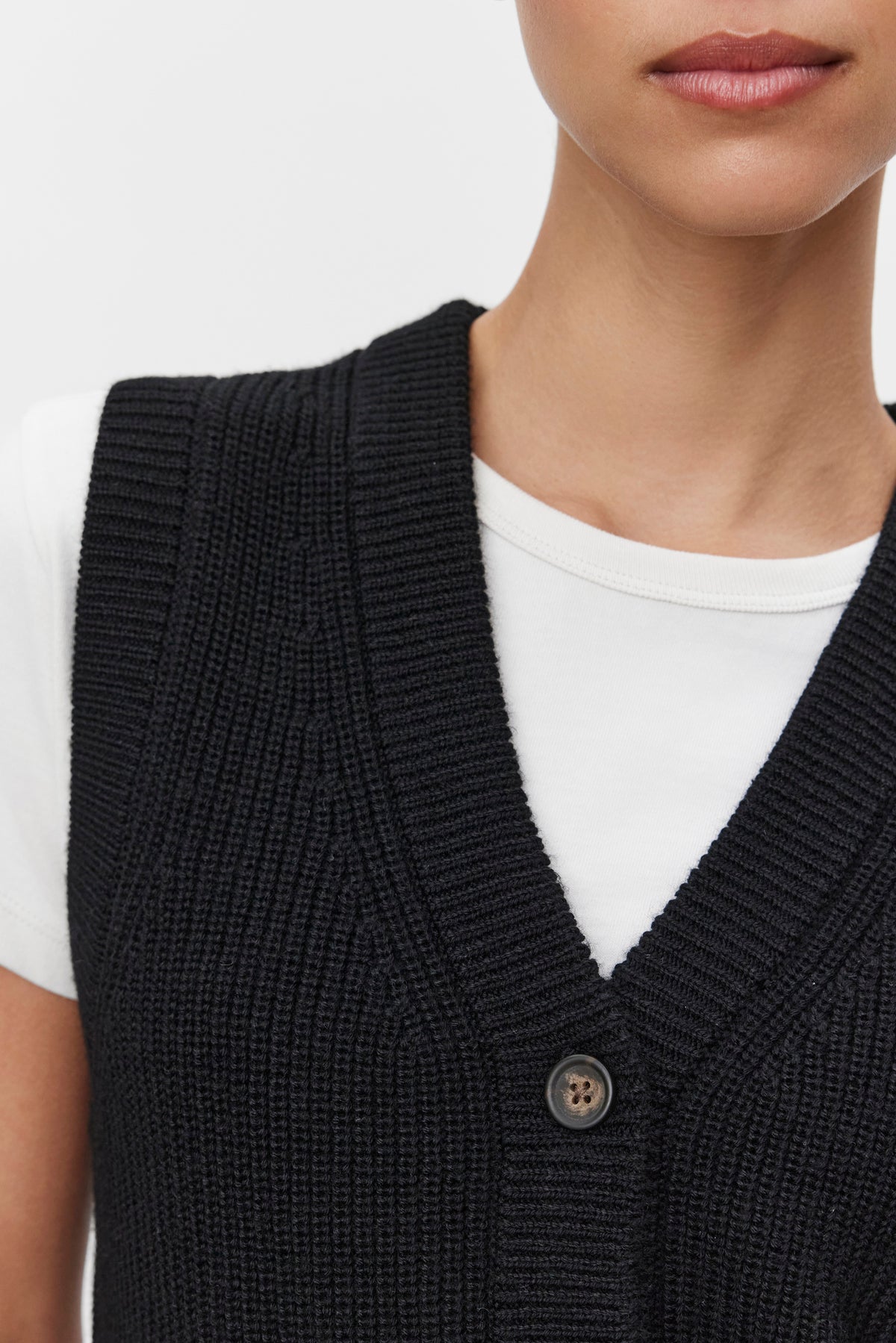   Close-up of a person wearing the LU SWEATER VEST by Velvet by Graham & Spencer, a black, rib-knit, buttoned sweater vest with a v-neckline, layered over a white t-shirt. Only the lower half of the face and upper torso are visible. 