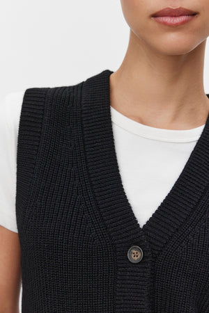 Close-up of a person wearing the LU SWEATER VEST by Velvet by Graham & Spencer, a black, rib-knit, buttoned sweater vest with a v-neckline, layered over a white t-shirt. Only the lower half of the face and upper torso are visible.