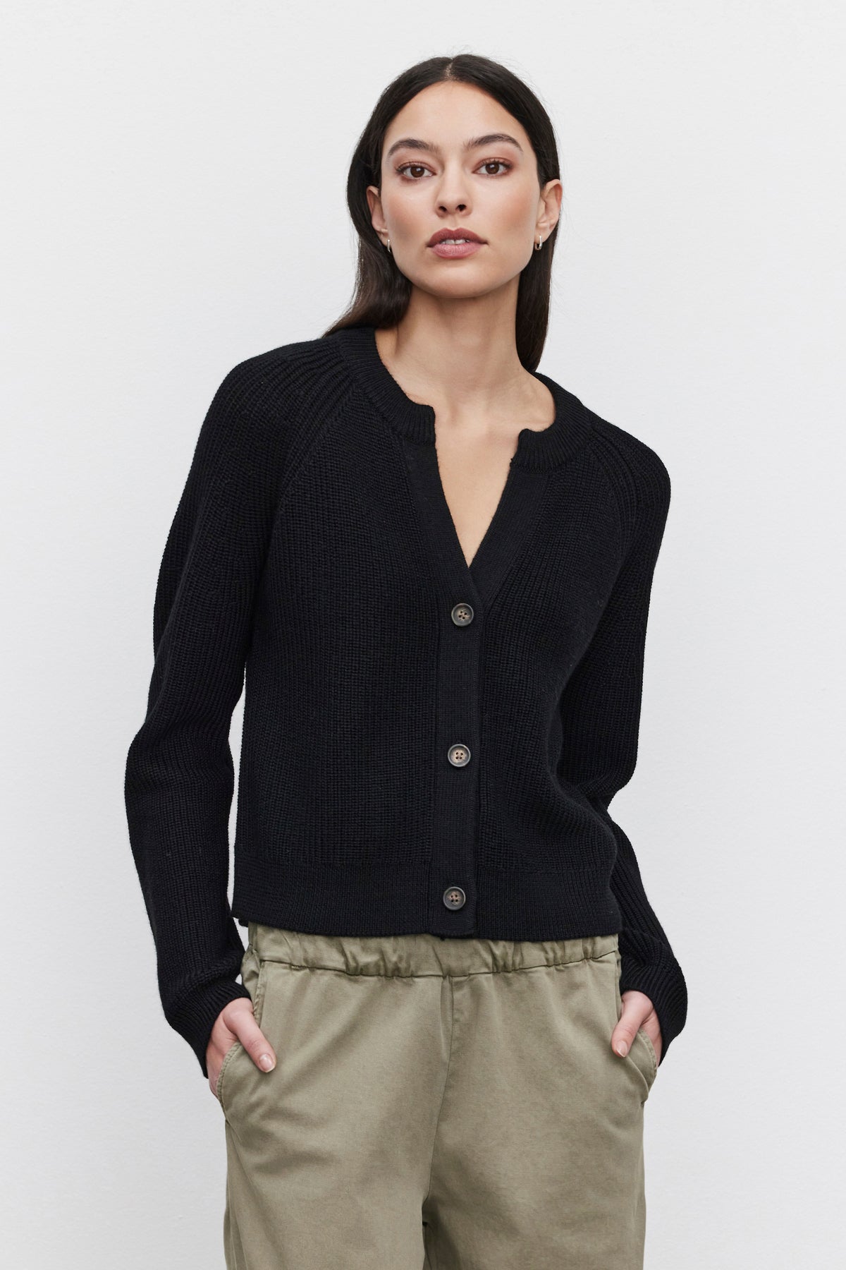   A woman with long dark hair is wearing the cozy SHAYLA CARDIGAN by Velvet by Graham & Spencer, paired with light khaki pants. Her hands are in her pockets, and she is standing against a plain white background. 