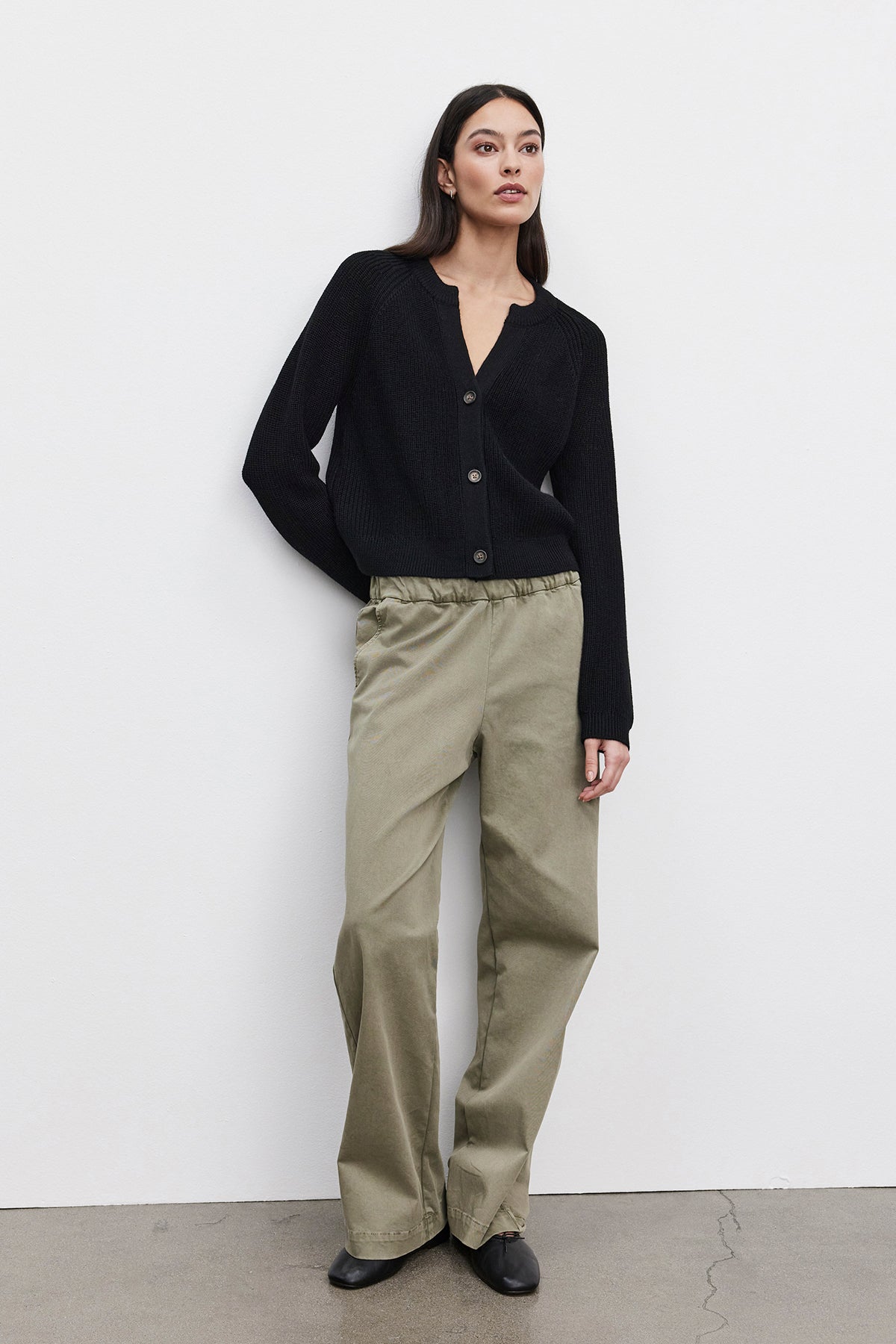   A person stands against a white wall wearing the SHAYLA CARDIGAN, a black wool blend button-up sweater by Velvet by Graham & Spencer, paired with olive green wide-legged pants. The individual's hands are in their pockets, and they are looking slightly to the right. 