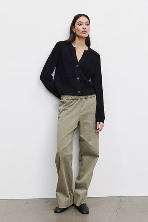 A person stands against a white wall wearing the SHAYLA CARDIGAN, a black wool blend button-up sweater by Velvet by Graham & Spencer, paired with olive green wide-legged pants. The individual's hands are in their pockets, and they are looking slightly to the right.