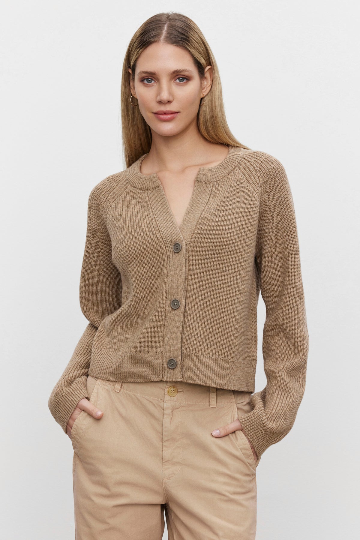   A woman with long blonde hair wears the SHAYLA CARDIGAN from Velvet by Graham & Spencer and beige pants, standing with her hands in her pockets against a neutral background. 