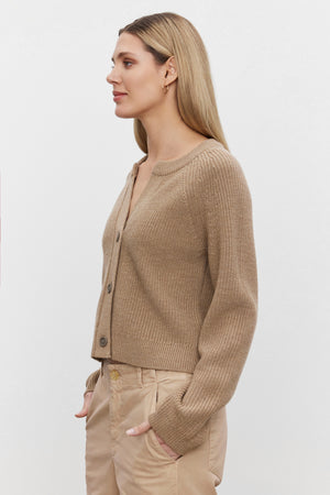 A woman with long blond hair, dressed in the SHAYLA CARDIGAN by Velvet by Graham & Spencer and tan pants, stands looking to the left against a plain white background.