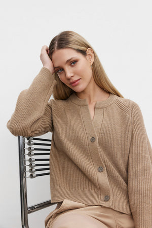 A blonde woman wearing the SHAYLA CARDIGAN by Velvet by Graham & Spencer, a cozy wool blend with a relaxed fit in beige, sits on a modern chair against a plain white background, her left hand resting on her head.