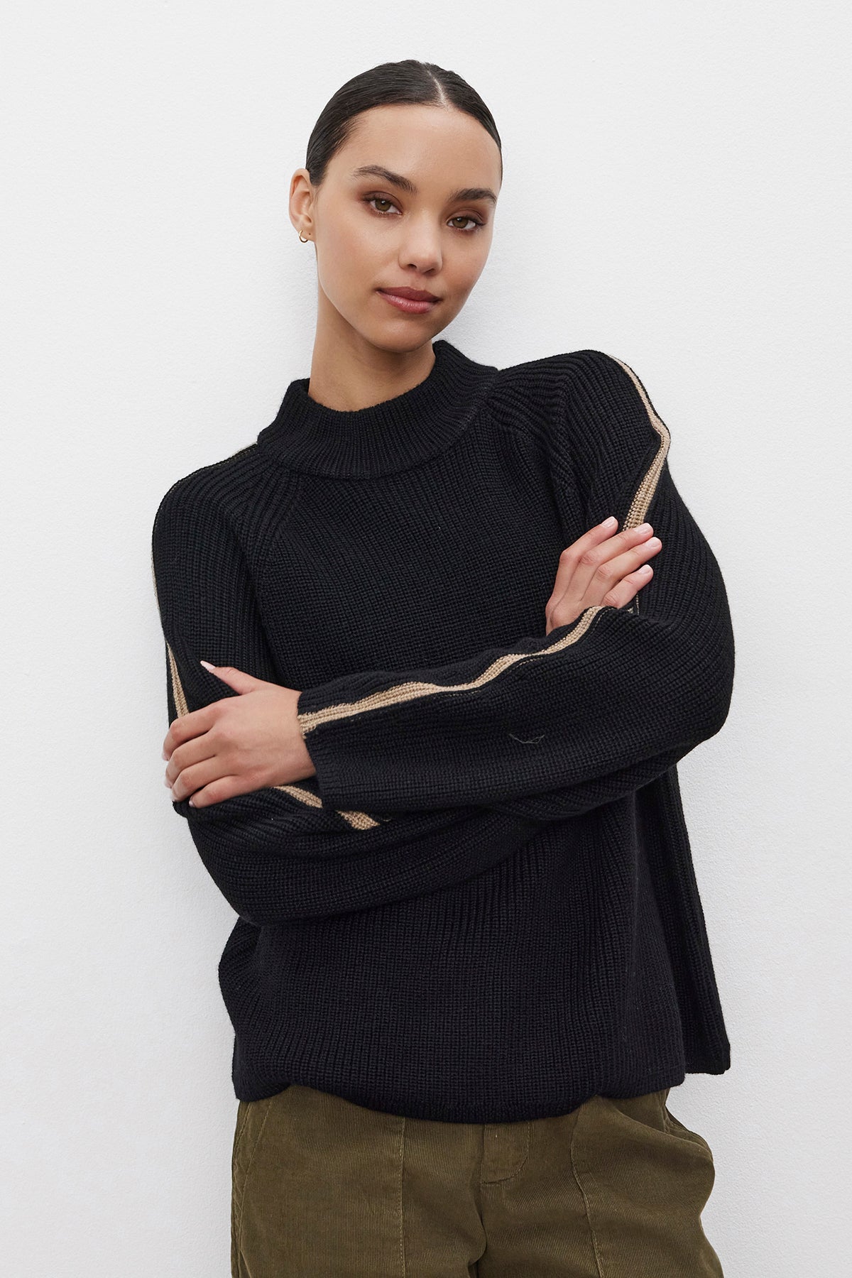   A person wearing the TEAGAN SWEATER by Velvet by Graham & Spencer, a black wool blend piece featuring light stripes on the sleeves, stands against a white background with their arms crossed. 