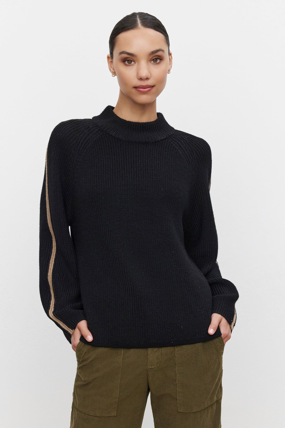   A person stands against a plain background wearing the cozy and comfortable Velvet by Graham & Spencer TEAGAN SWEATER, a black long-sleeve wool blend with a tan stripe on the sleeve, accompanied by olive green pants. 