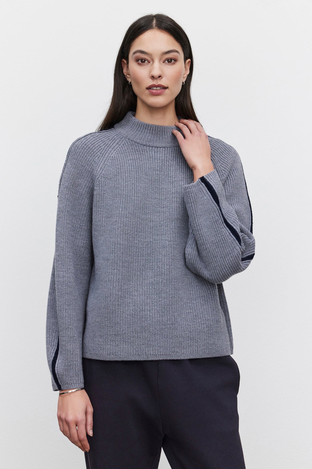   A person with long dark hair is wearing the TEAGAN SWEATER by Velvet by Graham & Spencer. The grey ribbed wool blend sweater, featuring dark trim on the sleeves, has a mock neck design and offers a relaxed silhouette. They are standing against a plain background. 