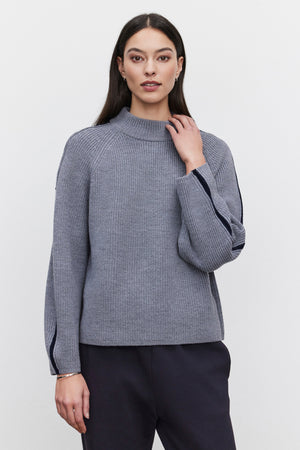 A person with long dark hair is wearing the TEAGAN SWEATER by Velvet by Graham & Spencer. The grey ribbed wool blend sweater, featuring dark trim on the sleeves, has a mock neck design and offers a relaxed silhouette. They are standing against a plain background.