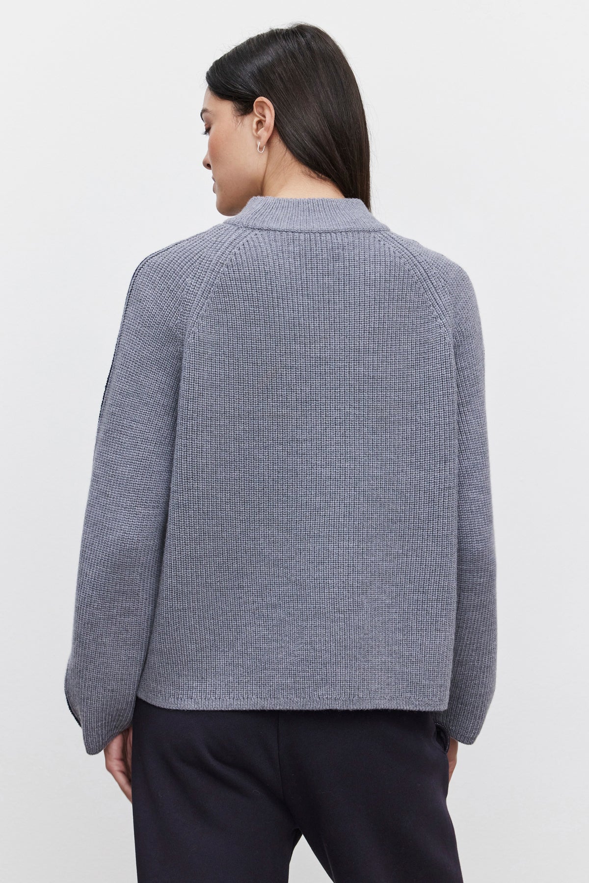   A person with long dark hair, facing away from the camera, is wearing the TEAGAN SWEATER by Velvet by Graham & Spencer, a grey ribbed wool blend sweater with a mock neck design, paired with dark pants. 