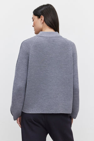 A person with long dark hair, facing away from the camera, is wearing the TEAGAN SWEATER by Velvet by Graham & Spencer, a grey ribbed wool blend sweater with a mock neck design, paired with dark pants.