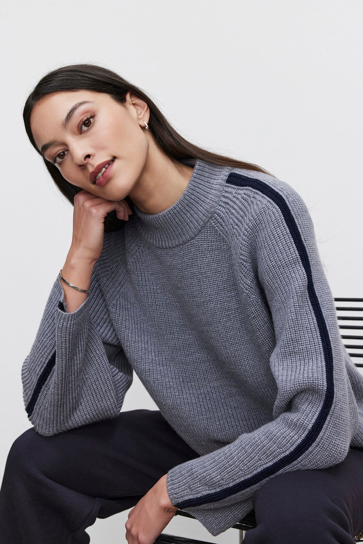   A woman wearing the TEAGAN SWEATER by Velvet by Graham & Spencer, characterized by its gray wool blend and dark striped sleeves, sits on a chair with a relaxed posture as she rests her head on her hand. 