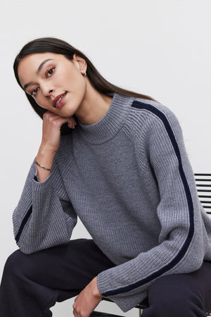 A woman wearing the TEAGAN SWEATER by Velvet by Graham & Spencer, characterized by its gray wool blend and dark striped sleeves, sits on a chair with a relaxed posture as she rests her head on her hand.