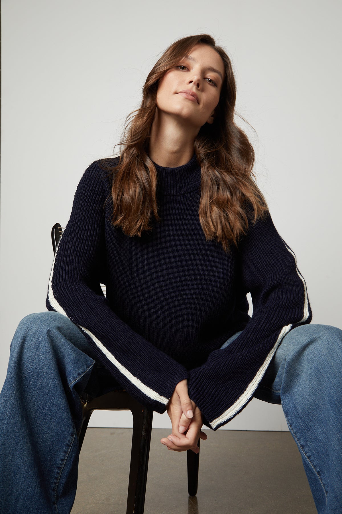   The model is sitting on a chair wearing cozy jeans and a Velvet by Graham & Spencer TEAGAN MOCK NECK SWEATER. 