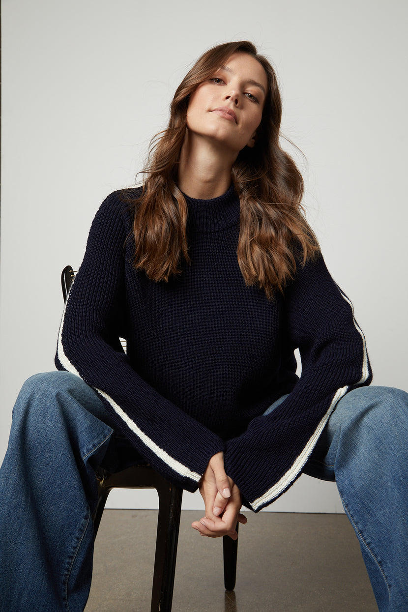 The model is sitting on a chair wearing cozy jeans and a Velvet by Graham & Spencer TEAGAN MOCK NECK SWEATER.