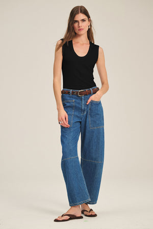 A woman stands against a plain background wearing the ESTINA TANK TOP by Velvet by Graham & Spencer, wide-leg blue jeans with front pockets, a brown belt, and brown sandals.