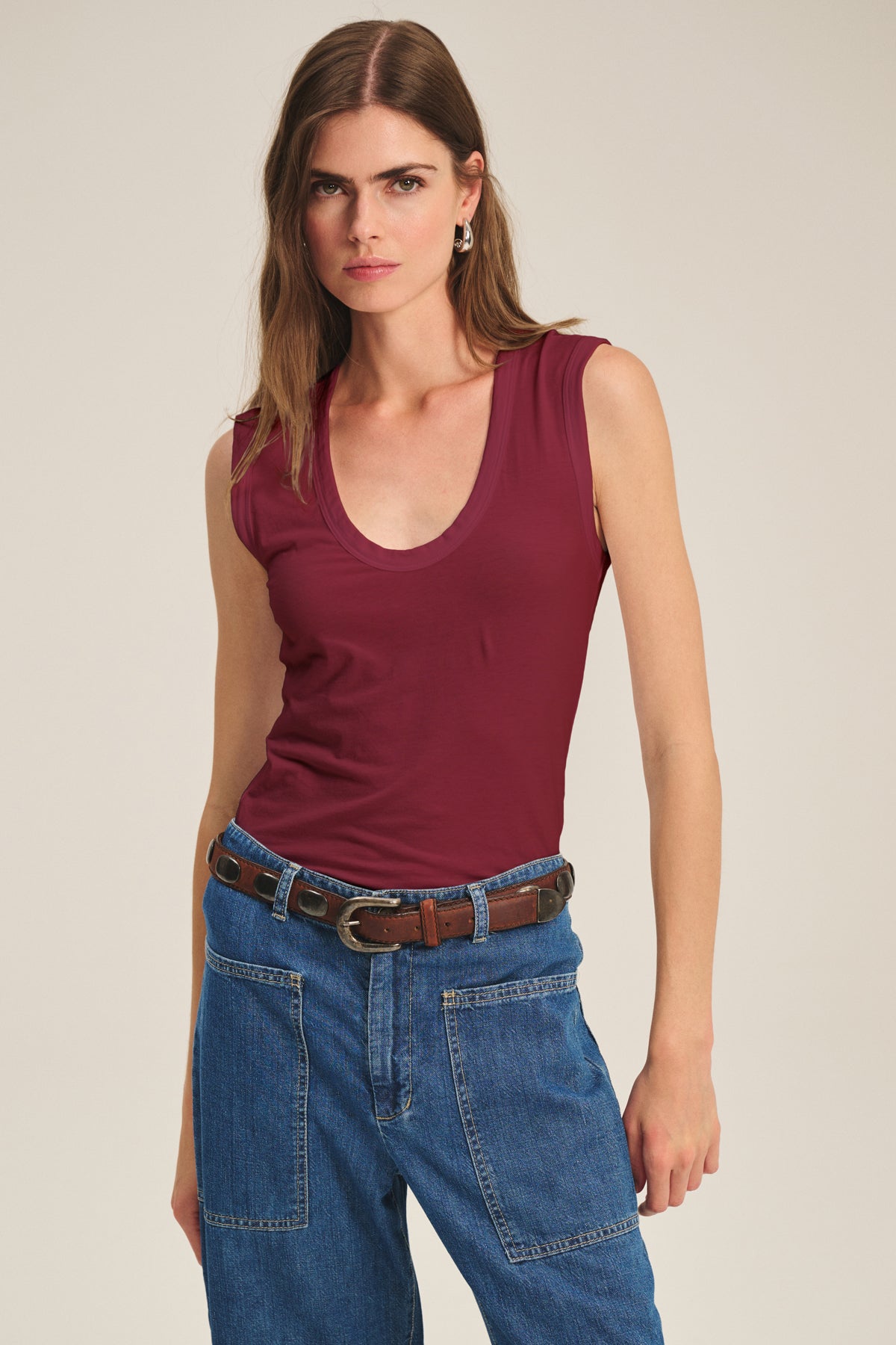  A woman wears the Velvet by Graham & Spencer ESTINA TANK TOP, a scoop-neck maroon tank, tucked into high-waisted blue jeans with a brown belt, standing against a plain background. 