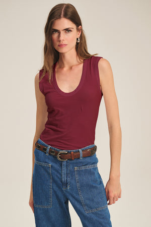 A woman wears the Velvet by Graham & Spencer ESTINA TANK TOP, a scoop-neck maroon tank, tucked into high-waisted blue jeans with a brown belt, standing against a plain background.