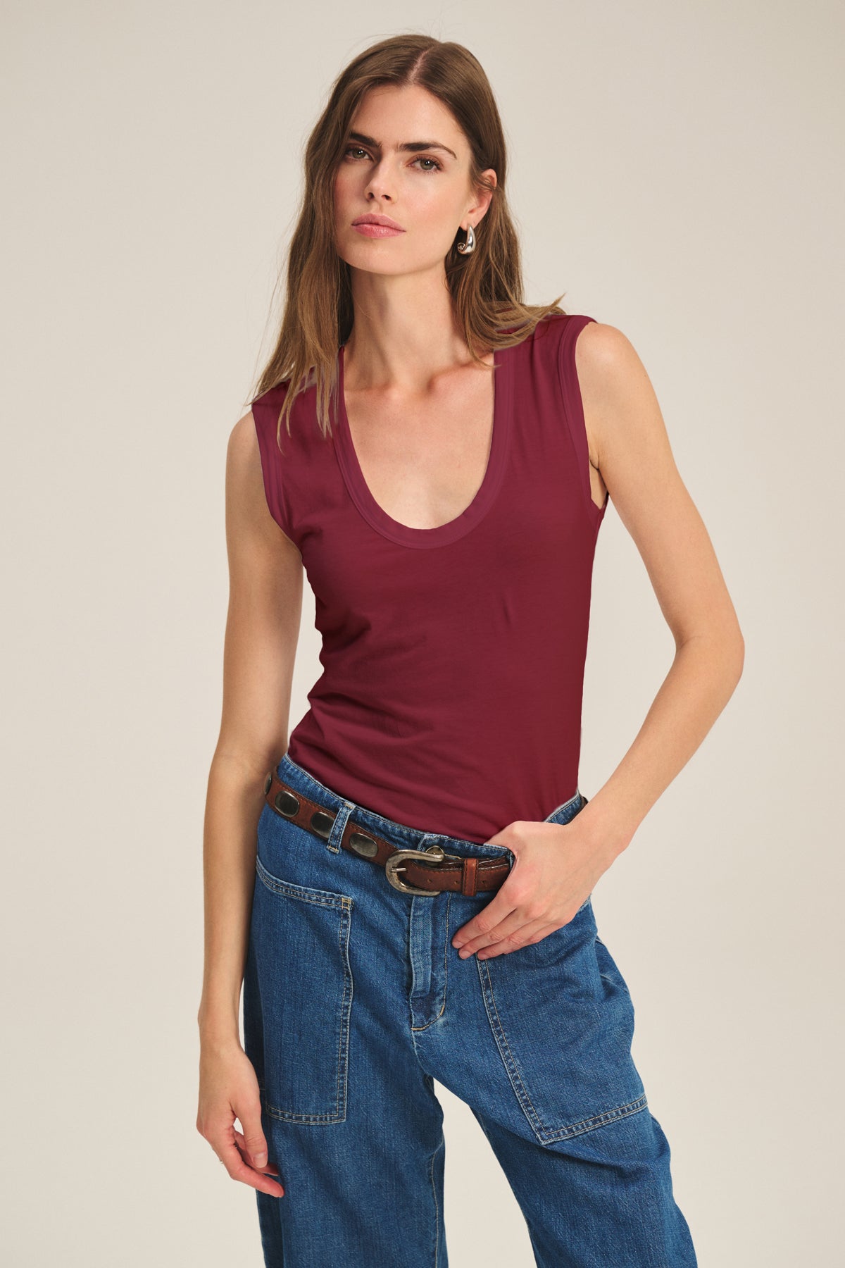   A woman wearing the ESTINA TANK TOP by Velvet by Graham & Spencer and blue jeans with a brown belt poses against a plain light background. 