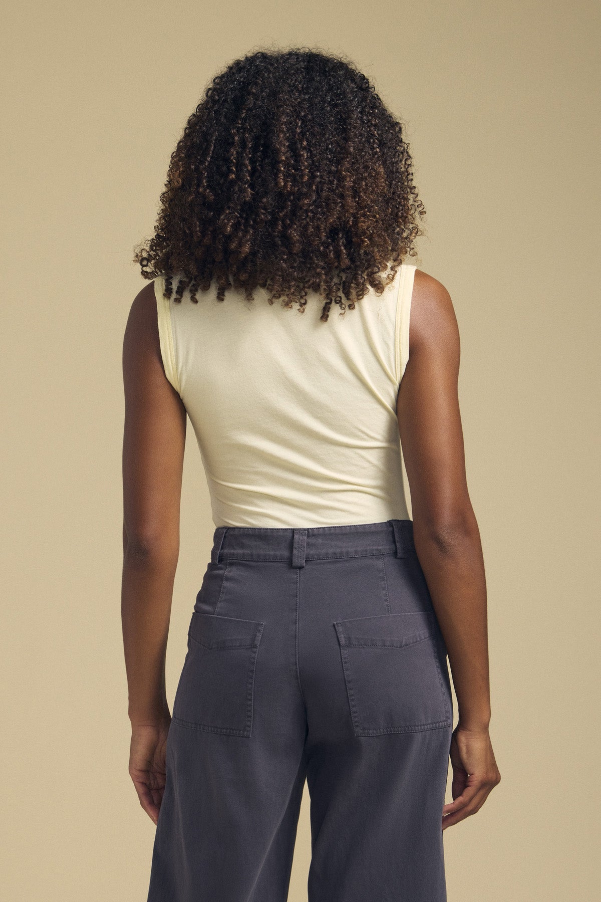 A person with curly hair seen from behind, wearing the laid-back ESTINA TANK TOP in light yellow by Velvet by Graham & Spencer and high-waisted gray pants with back pockets, standing against a beige background.-41929510191297