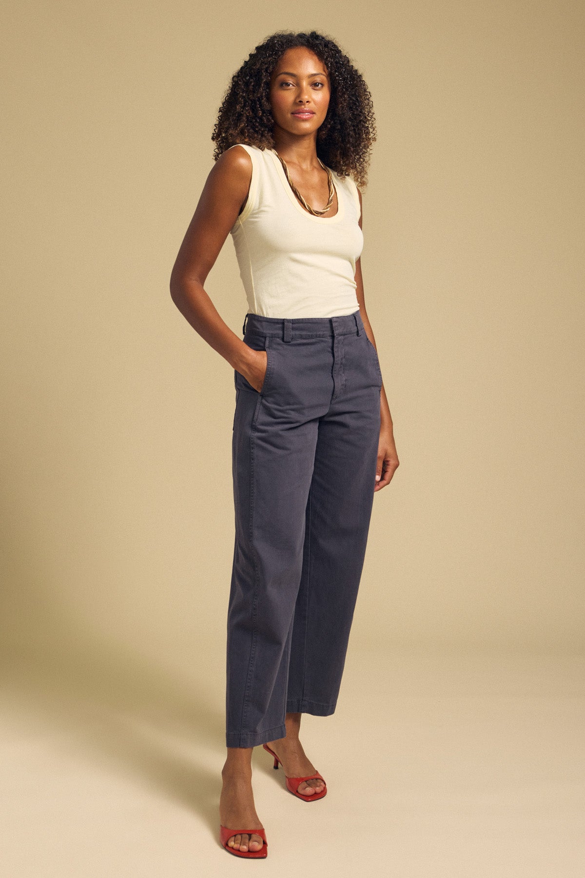 A woman stands with her hands in her pockets, wearing the ESTINA TANK TOP by Velvet by Graham & Spencer—a cream scoop-neck sleeveless top—paired with high-waisted gray pants, red heels, and a gold necklace. The look exudes effortless style against a beige background.