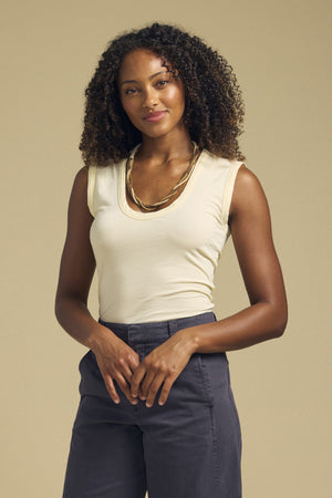 A woman with curly hair, styled in a gold necklace and gray pants, stands before a plain beige background wearing the ESTINA TANK TOP—an effortlessly chic sleeveless beige scoop-neck from Velvet by Graham & Spencer.