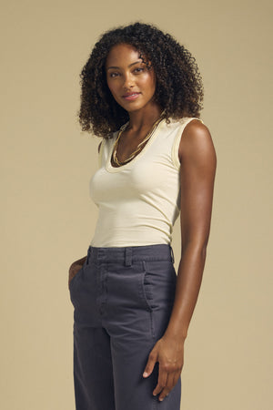 Woman with curly hair wears the Velvet by Graham & Spencer ESTINA TANK TOP—a scoop-neck, laid-back tank—paired with gray high-waisted pants as she stands against a plain beige background, exuding effortless style in gauzy whisper fabrics.