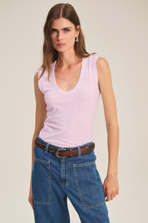 A woman wears the Velvet by Graham & Spencer ESTINA TANK TOP in a soft gauzy pink, paired with blue jeans with large front pockets and a brown belt, standing against a plain background.