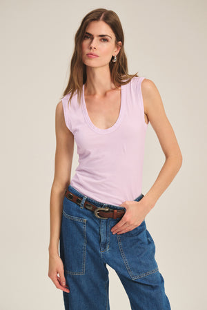 A woman wears the ESTINA TANK TOP by Velvet by Graham & Spencer in light pink, paired with blue jeans and a brown belt, standing against a plain background.