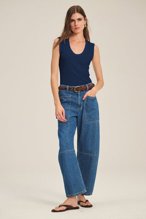 A woman wears the ESTINA TANK TOP by Velvet by Graham & Spencer, paired with wide-leg blue jeans, a brown belt, and brown sandals against a plain background.