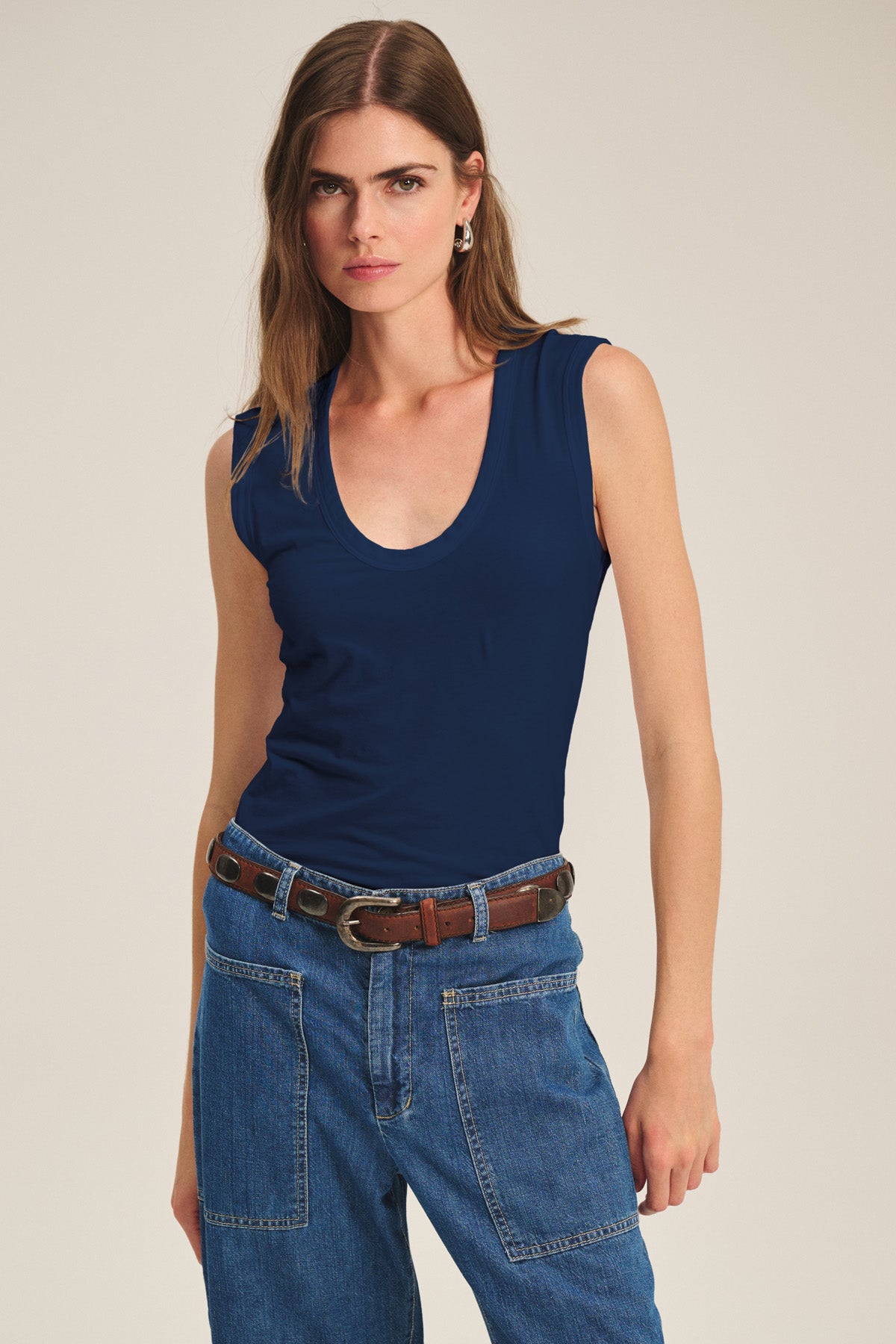 A woman wears the ESTINA TANK TOP by Velvet by Graham & Spencer, featuring a low scoop neck and sleeveless navy blue design, paired with high-waisted blue jeans and a brown belt, standing against a plain background.-40475799584961