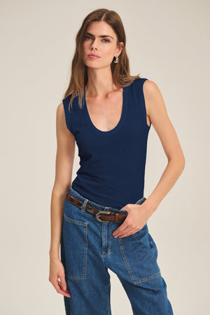 A woman stands against a plain background wearing the Velvet by Graham & Spencer ESTINA TANK TOP in navy blue, paired with blue jeans, a brown belt, and hoop earrings—her soft gauzy tank adds an effortless touch to her look.
