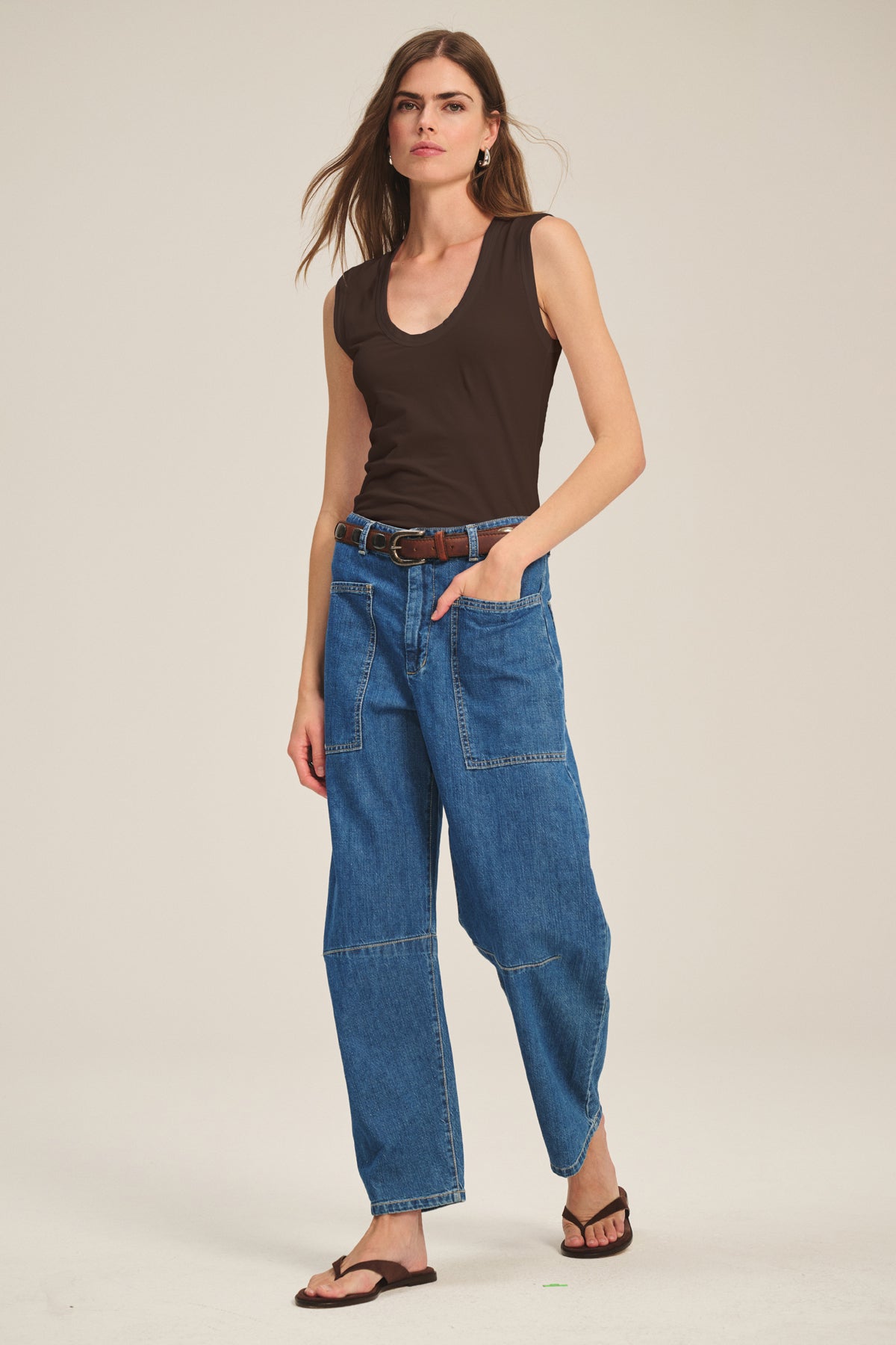   A woman stands against a plain background wearing the ESTINA TANK TOP by Velvet by Graham & Spencer, paired with blue patchwork jeans, a brown belt, and black sandals. 
