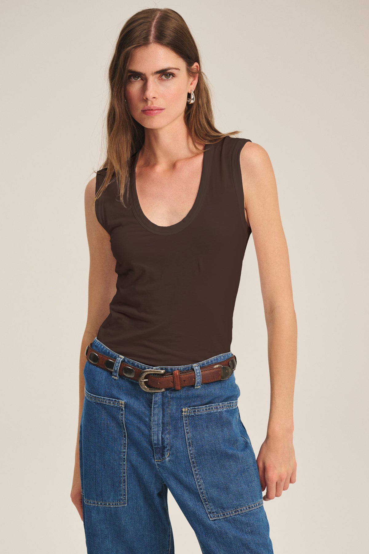 A woman stands against a plain background wearing the ESTINA TANK TOP by Velvet by Graham & Spencer, paired with blue jeans featuring patch pockets and a brown belt.-40182511894721