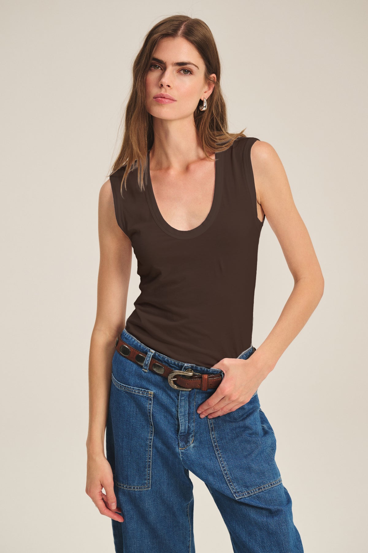   A woman wearing the ESTINA TANK TOP by Velvet by Graham & Spencer in dark brown with blue jeans stands against a plain beige background, looking at the camera. 
