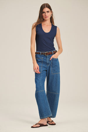 A woman wears the ESTINA TANK TOP by Velvet by Graham & Spencer—navy, gauzy, and sleeveless—paired with wide-leg blue jeans, a brown belt, and brown sandals against a plain background.
