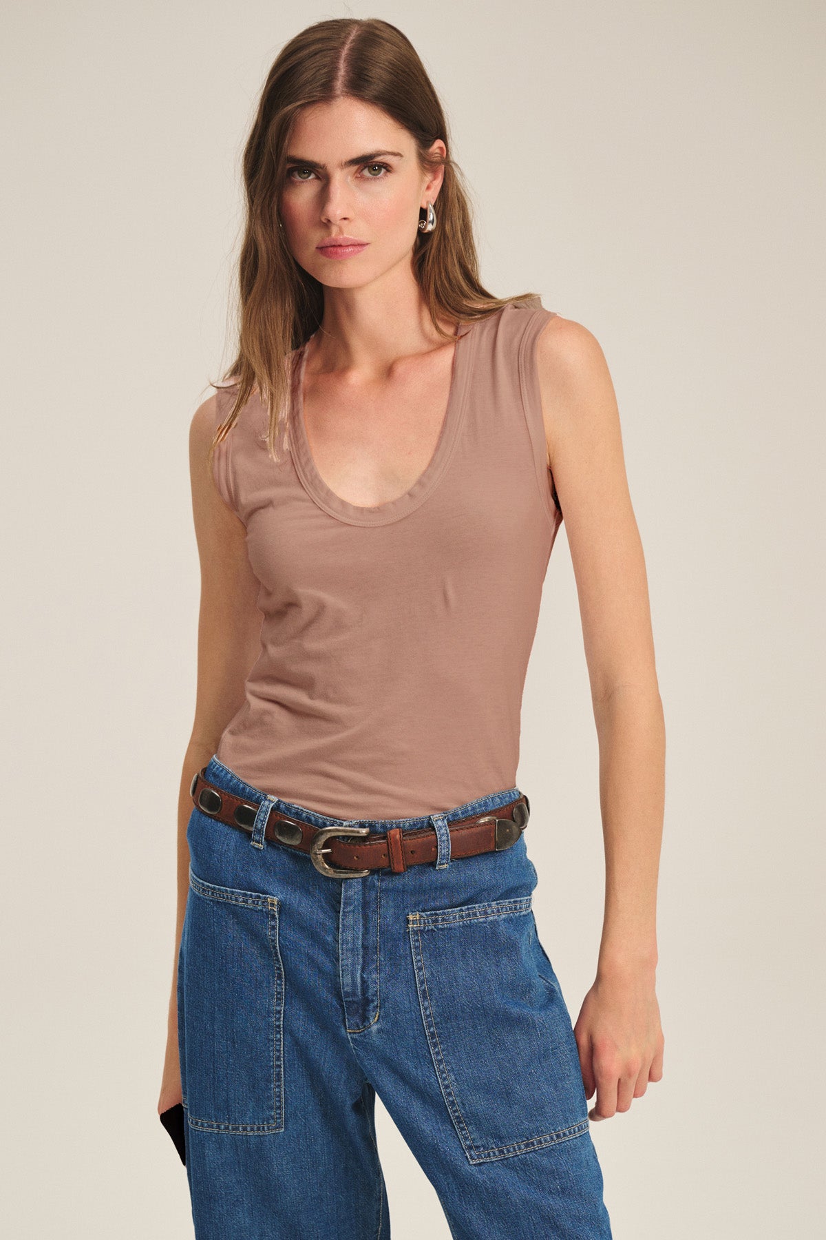 A woman wears the Velvet by Graham & Spencer ESTINA TANK TOP, a sleeveless beige scoop-neck tank, paired with blue jeans and a brown belt against a plain background.-41637036556481