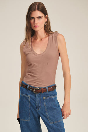 A woman wears the Velvet by Graham & Spencer ESTINA TANK TOP, a sleeveless beige scoop-neck tank, paired with blue jeans and a brown belt against a plain background.