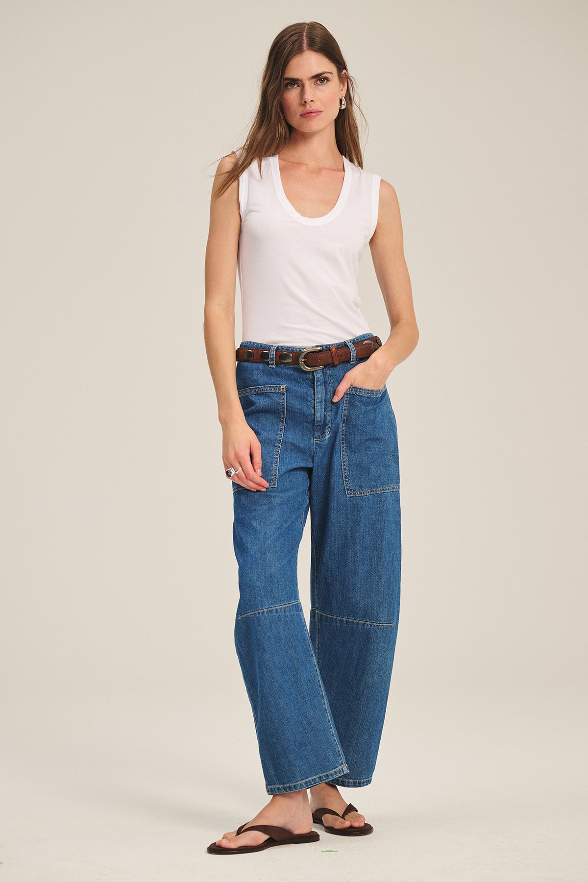 A woman stands in a studio wearing the ESTINA TANK TOP by Velvet by Graham & Spencer, styled with wide-leg blue jeans with front pockets, a brown belt, and brown sandals.-39600694493377