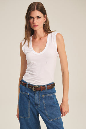 A woman in a Velvet by Graham & Spencer ESTINA Tank Top, blue jeans with large front pockets, and a brown belt stands against a plain background.