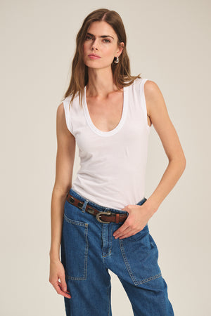 A woman wearing the ESTINA TANK TOP by Velvet by Graham & Spencer in white, paired with blue jeans and a brown belt, stands against a plain background.