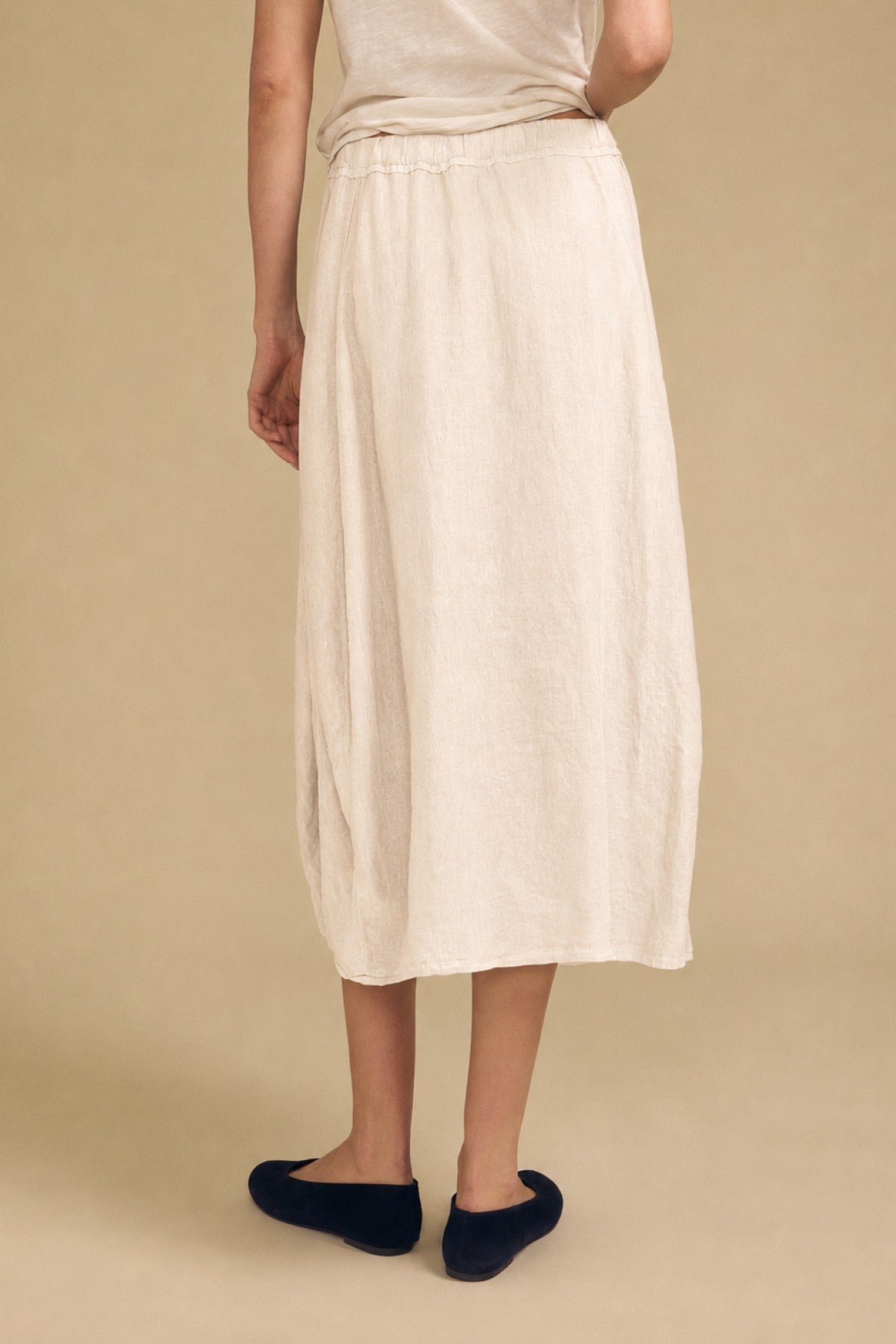 A person is seen from behind, standing against a plain beige background, wearing Velvet by Graham & Spencer’s FAE LINEN SKIRT in light beige—an ankle-length skirt featuring an elastic waistband—paired with black shoes.-42003448824001