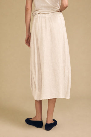 A person is seen from behind, standing against a plain beige background, wearing Velvet by Graham & Spencer’s FAE LINEN SKIRT in light beige—an ankle-length skirt featuring an elastic waistband—paired with black shoes.