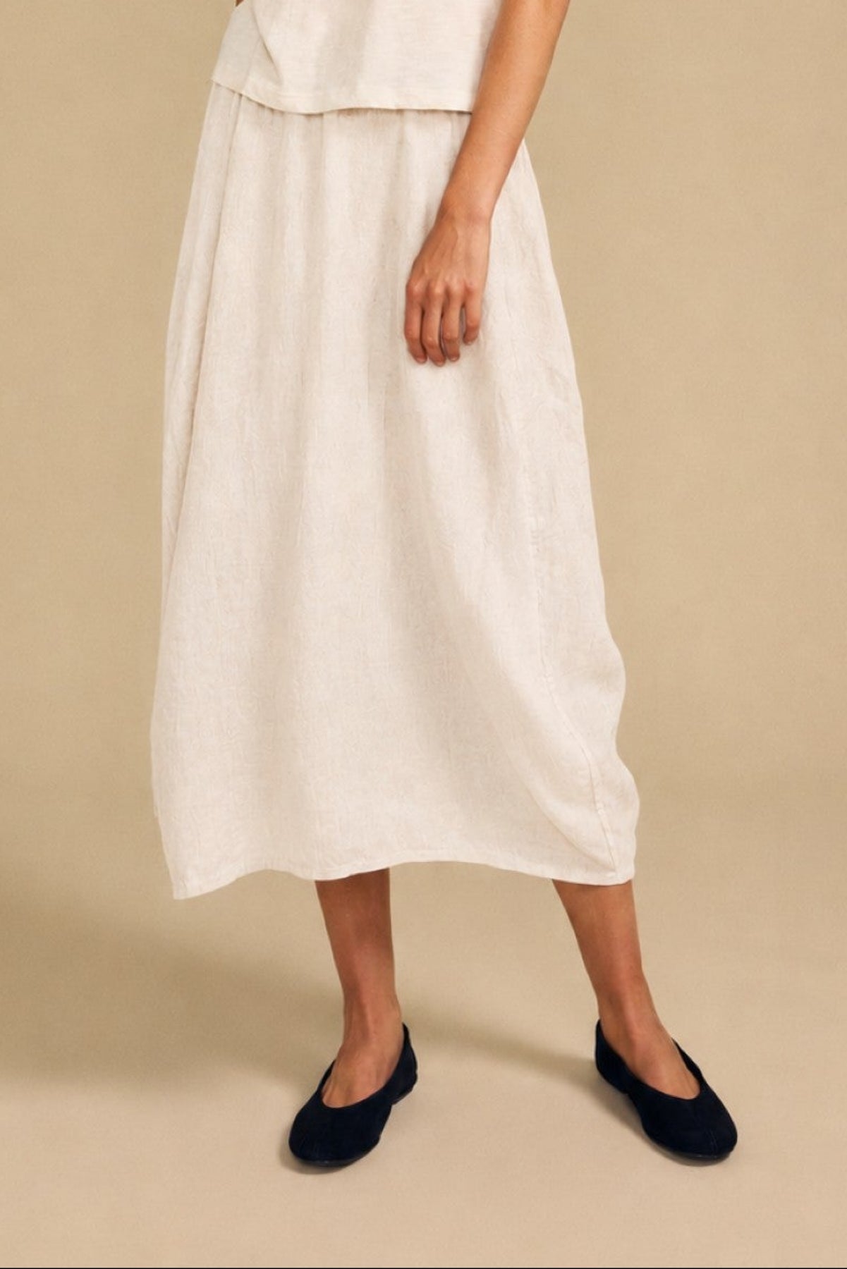 A woman stands against a beige background wearing a short-sleeve cream top and the FAE LINEN SKIRT by Velvet by Graham & Spencer—a matching linen maxi skirt featuring an elastic waistband for a relaxed A-line silhouette—paired with black flats.