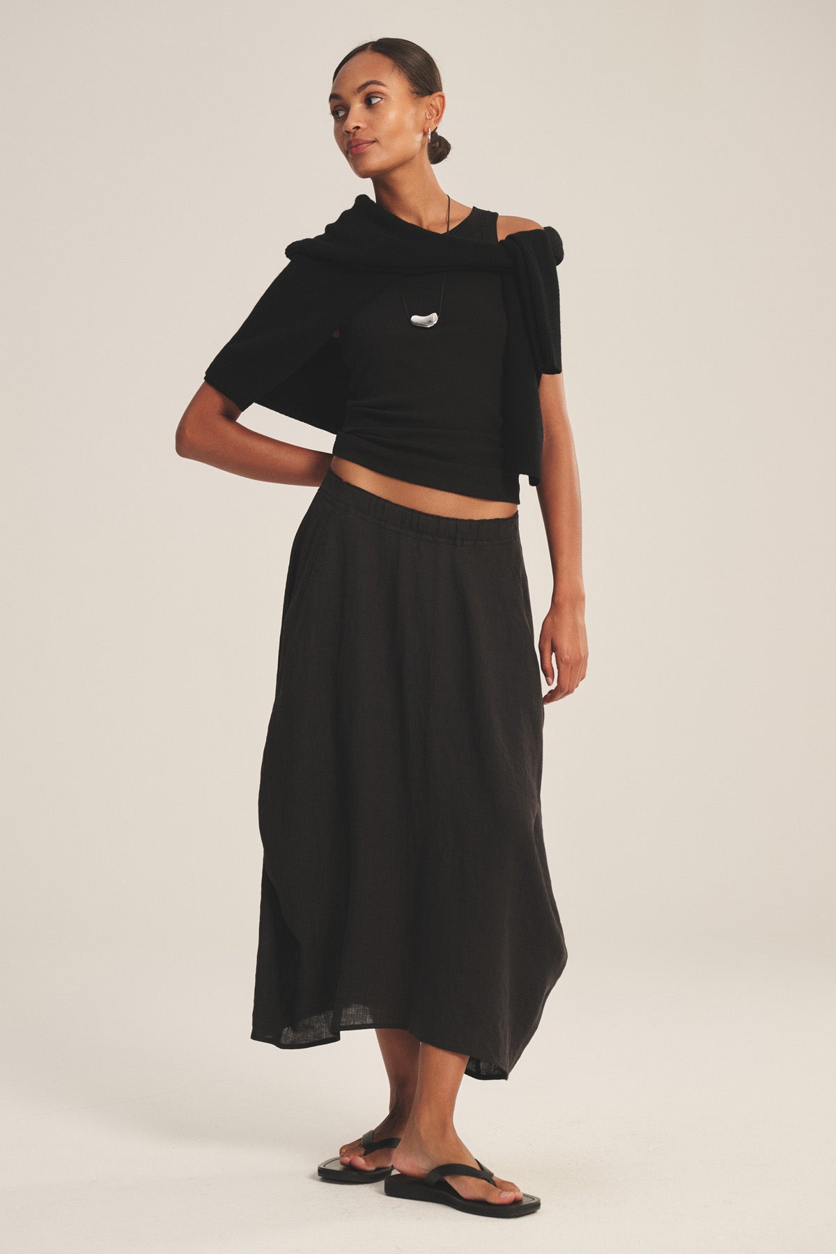 A woman stands against a plain background wearing a black off-shoulder top, black sandals, and the FAE LINEN SKIRT by Velvet by Graham & Spencer—a spring-weight A-line skirt with an elastic waistband—plus a black sweater draped over her shoulders.-40733683613889