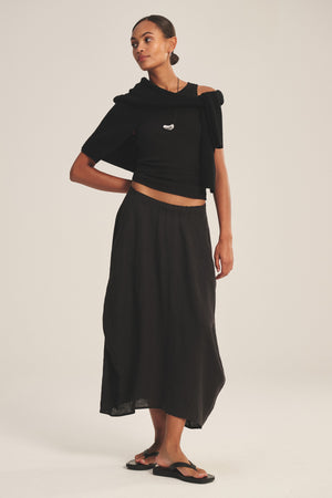 A woman stands against a plain background wearing a black off-shoulder top, black sandals, and the FAE LINEN SKIRT by Velvet by Graham & Spencer—a spring-weight A-line skirt with an elastic waistband—plus a black sweater draped over her shoulders.