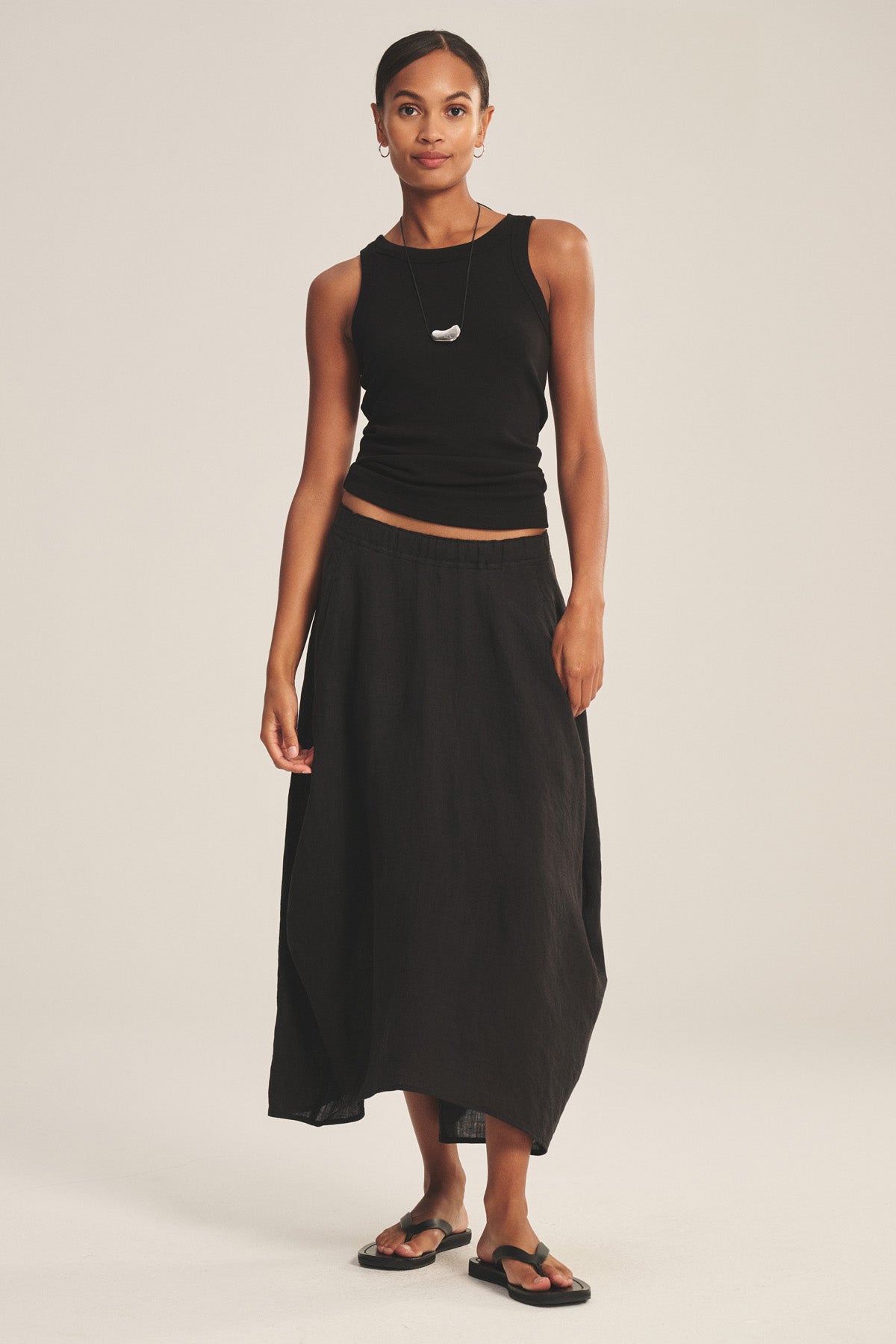 A woman stands against a plain background wearing a black spring-weight linen sleeveless top, the Velvet by Graham & Spencer FAE LINEN SKIRT with an elastic waistband, black sandals, and a necklace.-40733683646657