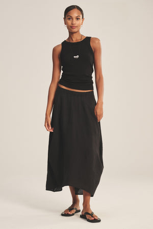 A woman stands against a plain background wearing a black spring-weight linen sleeveless top, the Velvet by Graham & Spencer FAE LINEN SKIRT with an elastic waistband, black sandals, and a necklace.