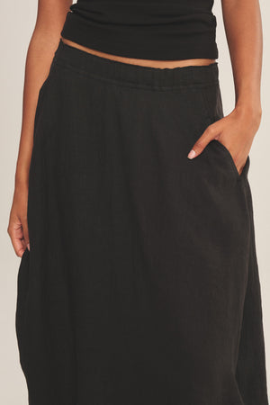 A person wears the FAE LINEN SKIRT by Velvet by Graham & Spencer—an A-line skirt with an elastic waistband and pockets—paired with a black sleeveless top, resting one hand casually in the skirt pocket.